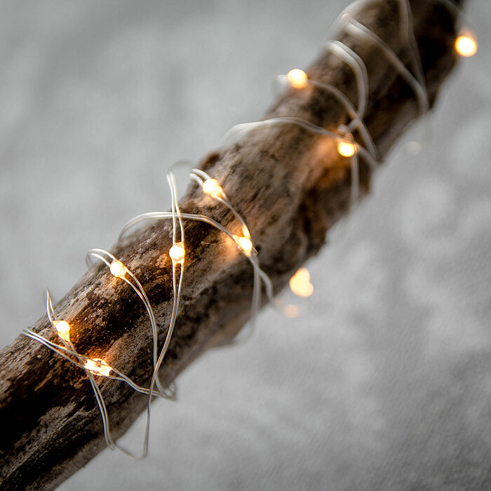 Ein altes Holzstück, das mit einer dünnen, silbernen Lichterkette umwickelt ist. Die kleinen, warmweißen Lichter der Kette funkeln dezent und geben dem Bild eine gemütliche und rustikale Stimmung. Der Hintergrund ist unscharf und in Grautönen gehalten, wa