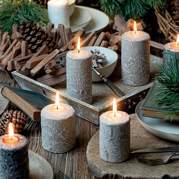 Sechs brennende Stumpenkerzen des Herstellers "Wenzel" in rustikaler Optik stehen auf einem Holztisch, umgeben von natürlichen Dekorationen wie Tannenzapfen und Kiefernnadeln. Die Szene vermittelt eine gemütliche, festliche Atmosphäre.