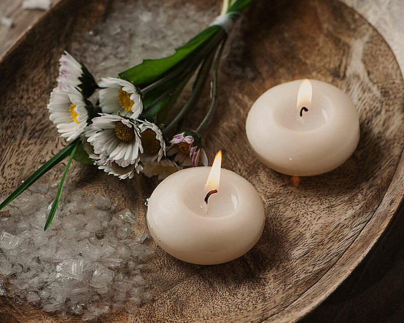 Eine rustikale Holzschale, mit Wasser und Kieselsand gefüllt, steht auf einem Holztisch. Auf der spiegelglatten Wasseroberfläche gleiten zwei brennende weiße Schwimmkerzen, ihr warmes Licht verbreitet eine behagliche Geborgenheit und eine tiefe Entspannung. Die Szenerie strahlt eine friedliche Stimmung aus, die zum Innehalten und Abschalten einlädt.