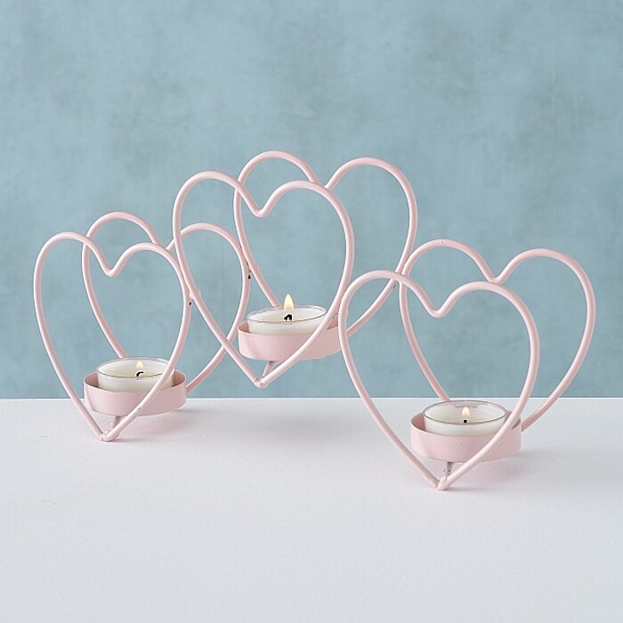 Drei brennende Teelichter sind in Pastellrosa Herz-förmigen Kerzenhaltern angeordnet. Die Herzen sind leicht voneinander versetzt, wodurch eine harmonische Anordnung entsteht. Der Hintergrund ist in einem sanften Hellblau gehalten, während die Teelichter 