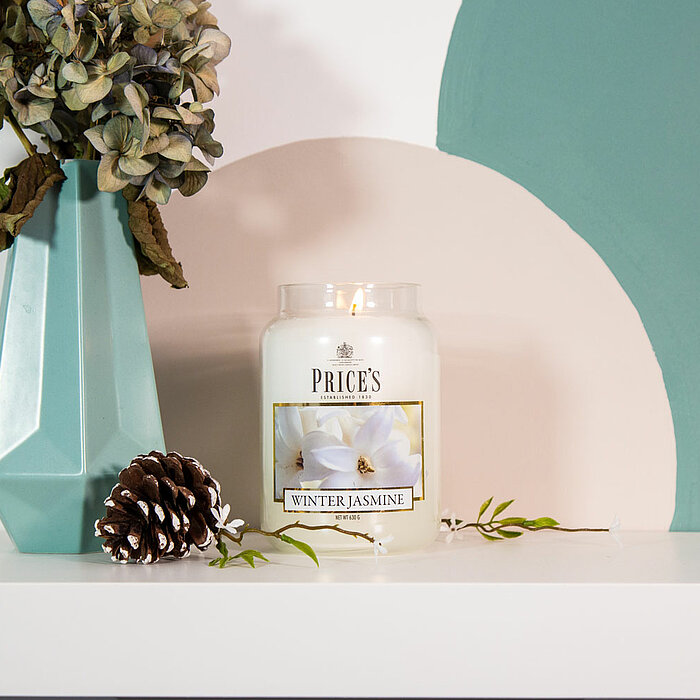 Eine brennende "Amber" Duftkerze des Herstellers Prices Candles steht auf einer hölzernen Unterlage. Daneben liegt ein Tannenzapfen und eine kleine getrocknete Blume in einer Vase. Im Hintergrund ist ein dunkelgraues Sofa mit gelben Kissen und einer Katze