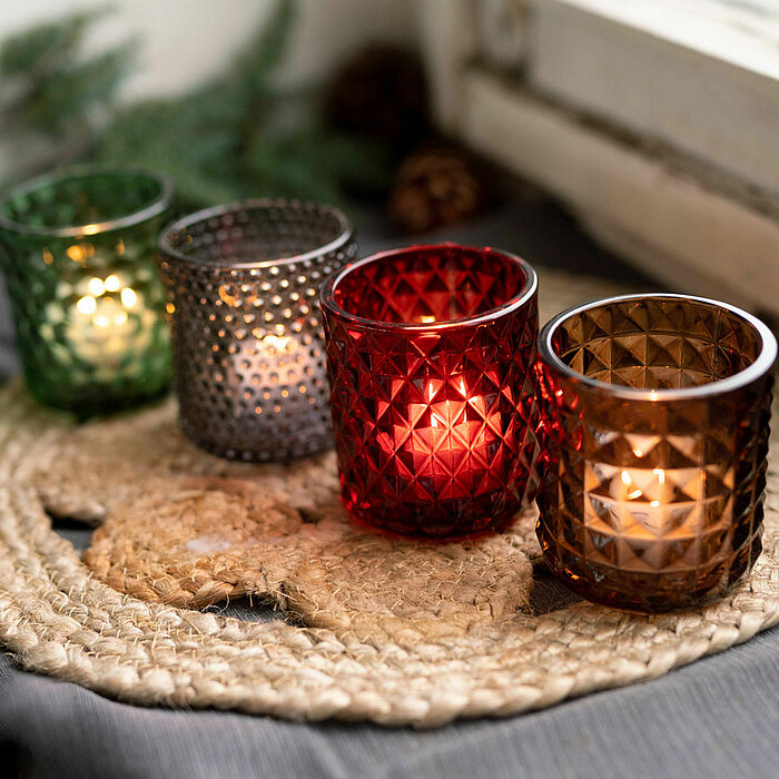 Vier brennende Teelichter von "Sandra Rich" in vielfältig farbigen, facettierten Glashaltern (Grün, Grau, Rot und Braun) stehen auf einer groben Jutematte. Im Hintergrund sind unscharf grüne Zweige sowie kleine Zapfen sichtbar, was eine gemütliche und nat