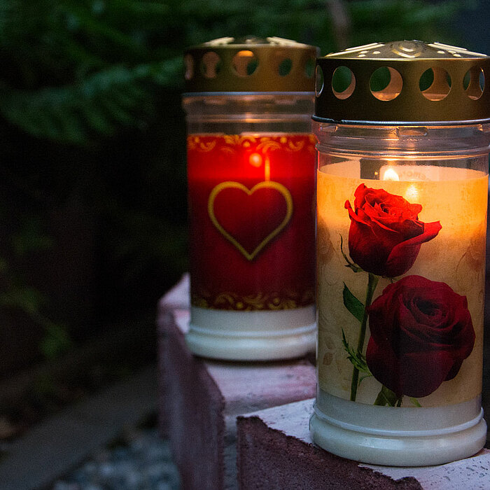 Zwei brennende Grabkerzen auf einem gemauerten Rand. Die linke Kerze ist rot und hat ein goldenes Herz als Dekoration. Die rechte Kerze ist mit einem Blumenmotiv von roten Rosen verziert. Im Hintergrund sind unscharfe, grüne Pflanzen zu sehen.