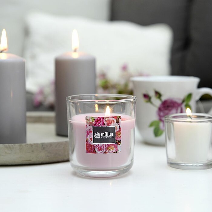 Fünf brennende Kerzen des Herstellers Müller, platziert auf einer runden, grauen Schale. Die Kerzen variieren in der Größe und Form, darunter zwei Glasbehälter-Kerzen, eine Stumpenkerze und zwei Teelichter. Im Hintergrund befindet sich ein graues Sofa mit