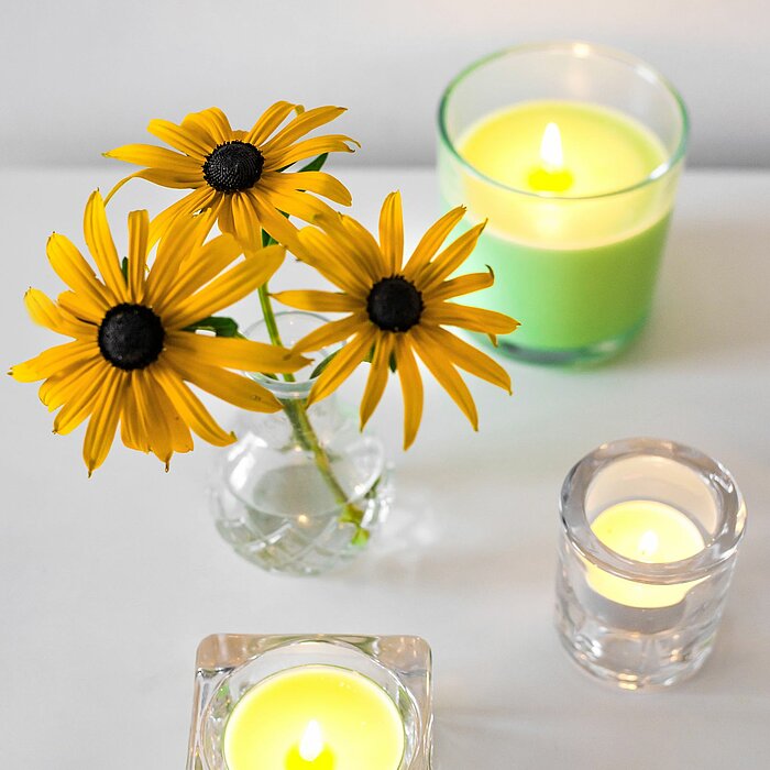 Vier brennende Bolsius Duftkerzen, angeordnet auf einer weißen Oberfläche. Eine der Kerzen ist in einem transparenten Glasbehälter mit hellgrünem Wachs. Die andere steht ebenfalls in einem Glasbehälter und enthält weißes Wachs. Zwei weitere Duftkerzen bef