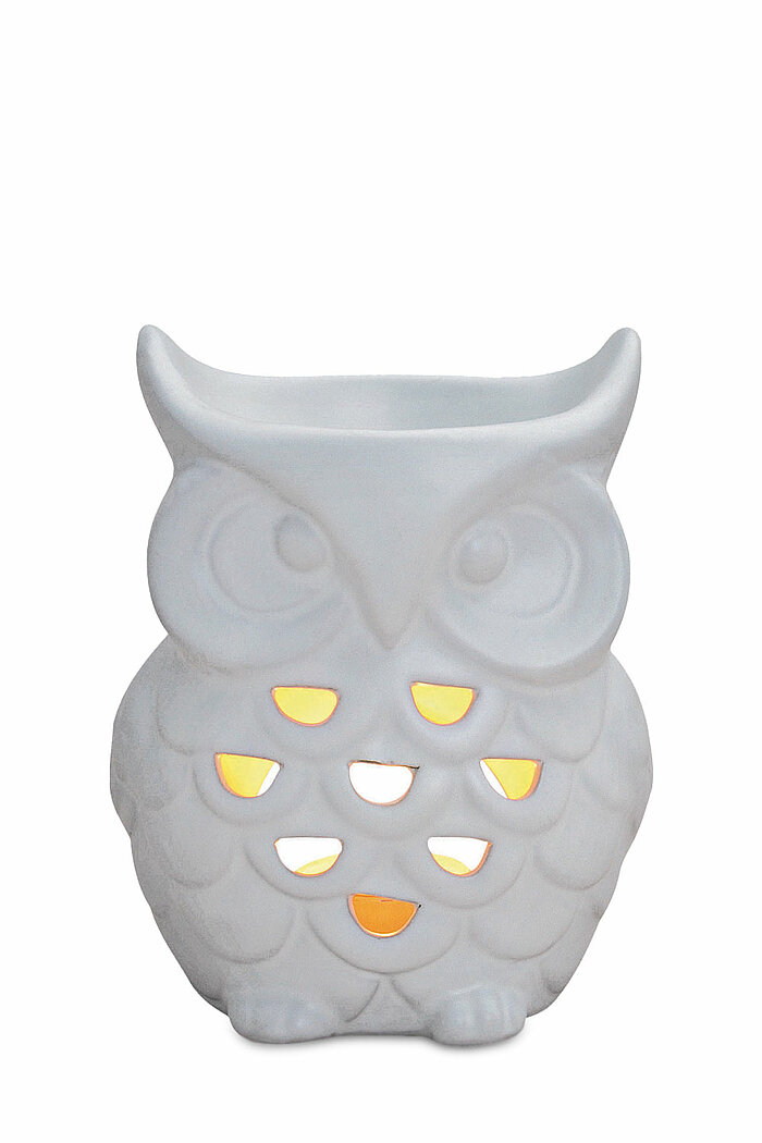 Ein weißer, eulenförmiger Teelichthalter aus Keramik. Die Eule hat große, runde Augen und ein detailliert ausgearbeitetes Federkleid, in dem mehrere herzförmige Löcher eingelassen sind. Durch diese Löcher scheint diffuses, warmes Licht, was dem Objekt ein