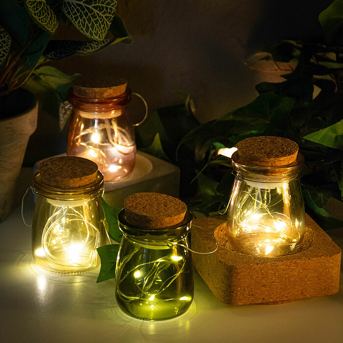 Vier kleine bauchige Glaslaternen mit Korkdeckeln stehen auf einem rustikalen Holzbrett. Jede Laterne enthält eine Lichterkette, die in warmen Gelbtönen leuchtet. Die Laternen haben verschiedene Farben: Rosa, Klar, Grün und Klar mit einem leicht goldenen 