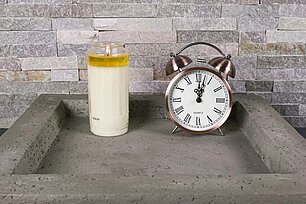In einer eckigen grauen Betonschale steht eine weisse große brennende Grabkerze aus reinem Pflanzenöl ohne Etikett der Marke Aeterna. Rechts neben der Grabkerze befindet sich ein schöner alter Wecker in Kupferfarbe, der die Uhrzeit kurz nach 0 Uhr anzeigt. Im Hintergrund sieht man eine steinerne Mauer.