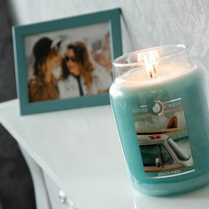Eine brennende Goose Creek Duftkerze in einem transparenten Glasbehälter mit der Aufschrift "Beach Vibes" steht auf einer weißen Kommode. Im Hintergrund befindet sich ein gerahmtes Foto von zwei Personen, das in einem türkisfarbenen Bilderrahmen steckt. D