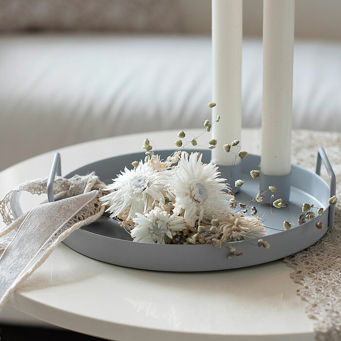 Zwei weiße, lange Kerzen von Halbach stehen in einem grauen Kerzenhalter auf einem weißen Tisch. Um die Kerzen herum sind getrocknete weiße Blüten und dekorative graue Stoffbänder arrangiert. Im Hintergrund ist ein helles Sofa zu sehen.