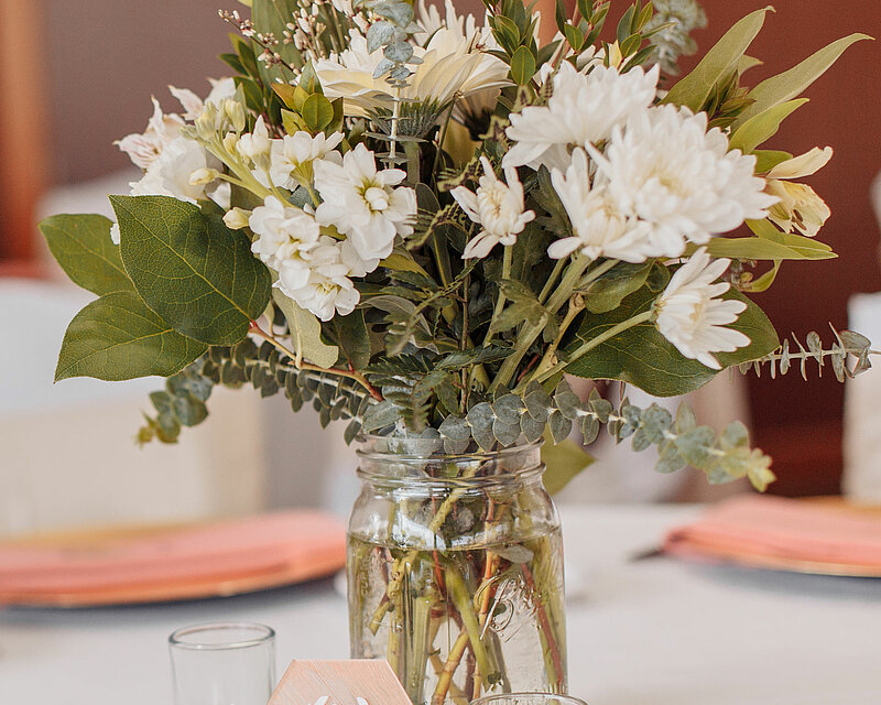 Auf einem weißen gedeckten Tisch steht eine runde Spiegelplatte von Sandra Rich. Auf der Spiegelplatte stehen zwei transparente Teelichthalter mit je einem Teelicht. Dazwischen steht eine große Glas Vase mit einem schönen Blumenstrauß mit weißen Blüten und grünen Blättern.