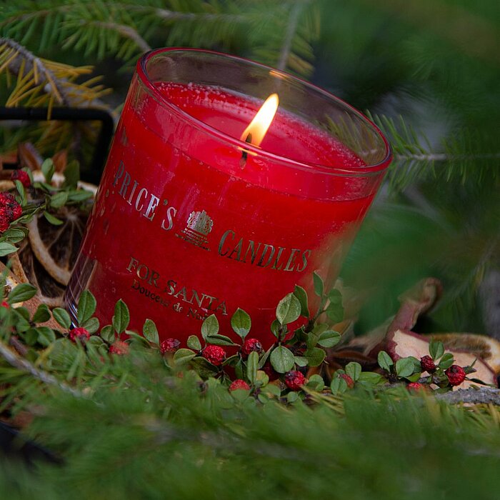 Eine brennende rote Duftkerze von "Price's Candles" in einem Glasbehälter, umgeben von grünen Zweigen und roten Beeren. Die goldene Inschrift auf dem Glas hebt das hochwertige Design hervor und gibt einen festlichen Touch. Im Vordergrund sind grüne Tannen