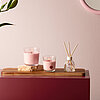 Ein harmonisch gestalteter Raum mit zwei rosa Bolsius Duftkerzen in Glasbehältern, die auf einem Holzbrett neben einem Glasgefäß mit Duftstäbchen stehen. Im Hintergrund ist eine zartrosa Wand mit einem runden Spiegel und einer dunkelroten Oberfläche sowie