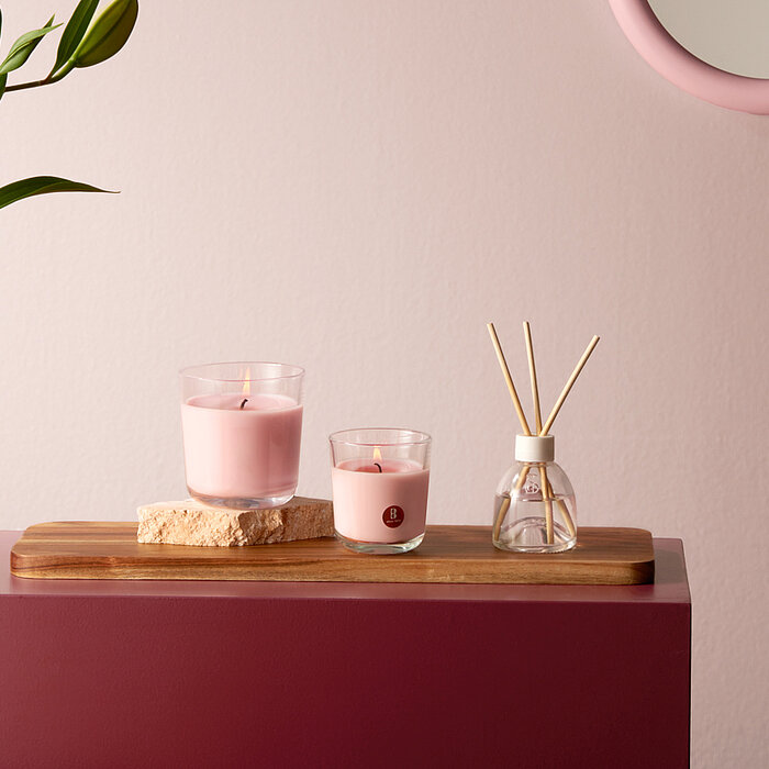 Ein harmonisch gestalteter Raum mit zwei rosa Bolsius Duftkerzen in Glasbehältern, die auf einem Holzbrett neben einem Glasgefäß mit Duftstäbchen stehen. Im Hintergrund ist eine zartrosa Wand mit einem runden Spiegel und einer dunkelroten Oberfläche sowie