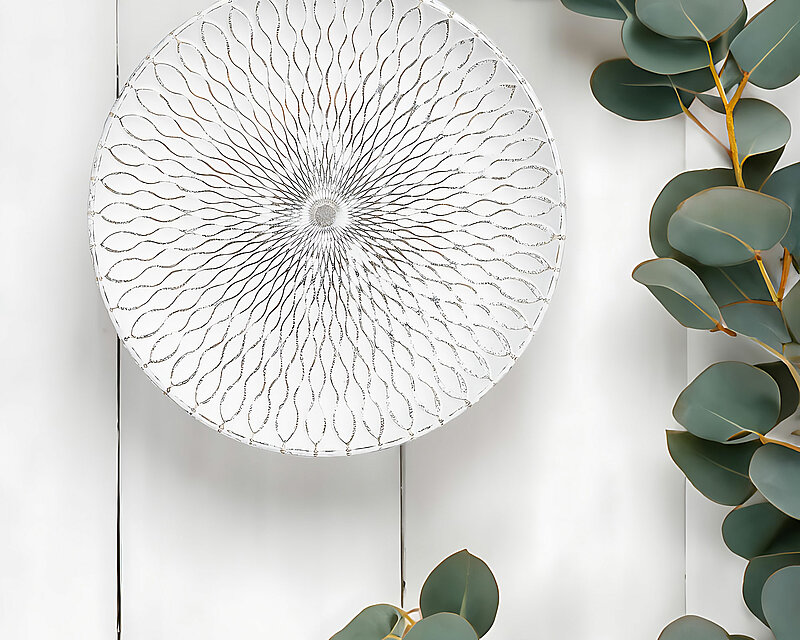 Man sieht frontal von oben auf einen weißen Holztisch. Am rechten Bildrand ragen einige Zweige einer Eukalyptuspflanze ins Bild. In der Mitte steht ein weißer Holzteller von Inge-Decor mit einer hübschen Ornamentverzierung.