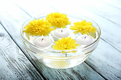 Das Bild zeigt eine Glasschale gefüllt mit Wasser und einigen schwimmenden gelben Blüten und drei brennenden weissen Schwimmkerzen. Die Farben des Bildes und die Schwimmkerzen verleihen dem Bild eine Sommerliche und frische Atmosphäre.