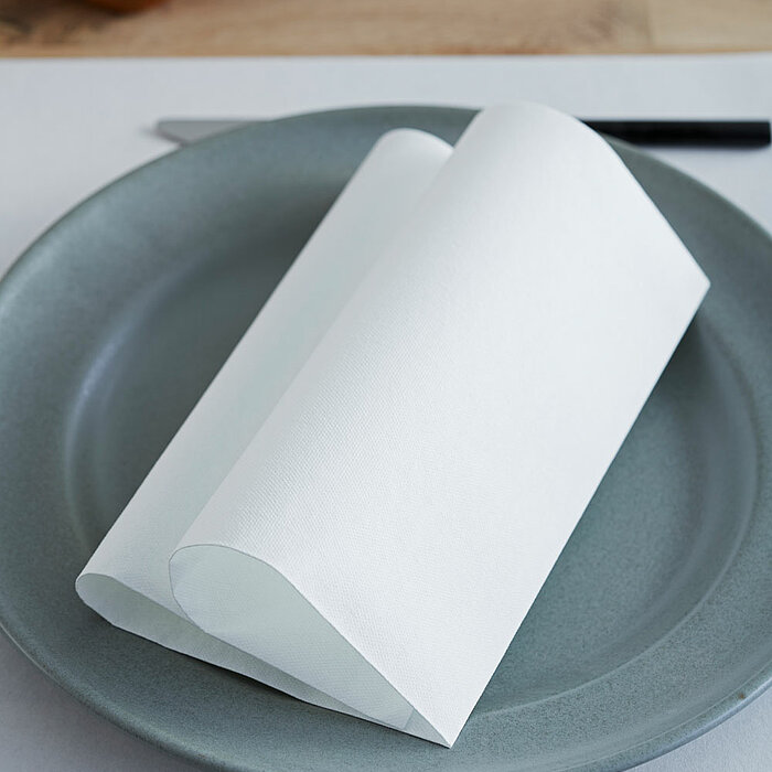 Ein grauer Teller mit sorgfältig gefalteten, weißen Servietten des Herstellers "Duni". Daneben liegen eine schwarze Gabel und ein Messer. Im Hintergrund befindet sich eine braune Ledertasche, die für ein stilvolles und gepflegtes Ambiente sorgt.