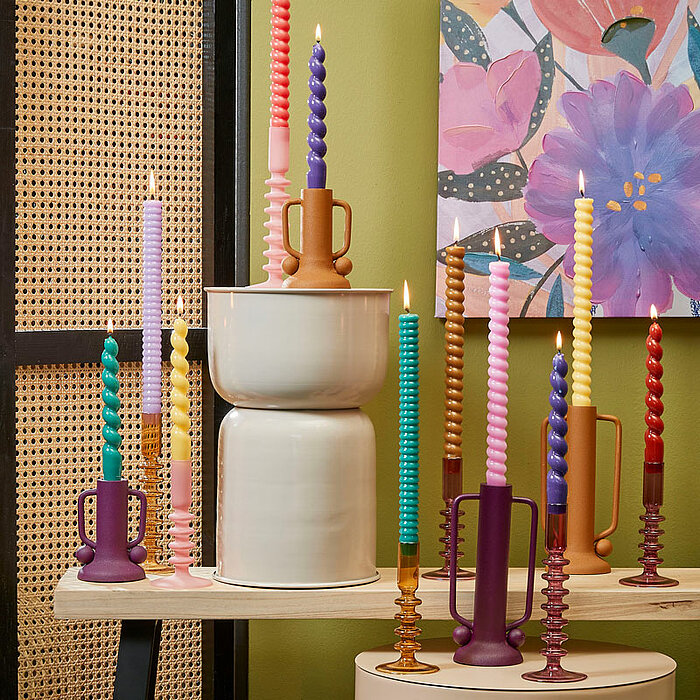 Eine lebendige Szenerie mit mehreren brennenden Kerzen vom Hersteller Edelman. Die Kerzen sind farbenfroh in hellblau, lila, rosa, gelb und weiteren Farben. Sie stehen auf verschieden hohen Kerzenhaltern und Sockeln, die ebenfalls farblich abgestimmt sind