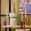 Eine lebendige Szenerie mit mehreren brennenden Kerzen vom Hersteller Edelman. Die Kerzen sind farbenfroh in hellblau, lila, rosa, gelb und weiteren Farben. Sie stehen auf verschieden hohen Kerzenhaltern und Sockeln, die ebenfalls farblich abgestimmt sind