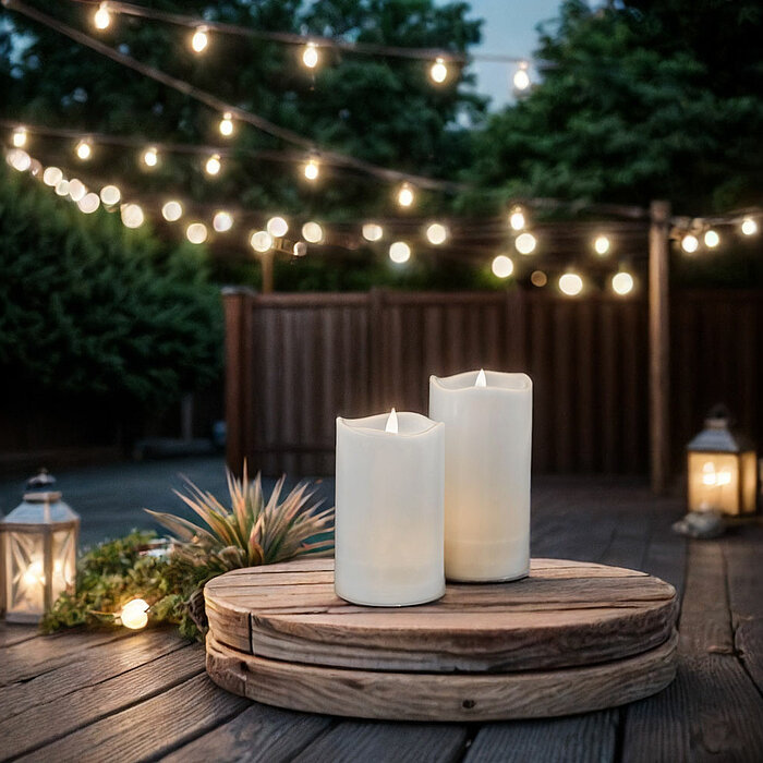 Zwei brennende weiße Stumpenkerzen stehen auf einem rustikalen Holztablett, das sich auf einer Holzterrasse befindet. Rundherum sind warme Lichterketten gespannt, die eine gemütliche und einladende Atmosphäre schaffen. Im Hintergrund stehen mehrere verzie