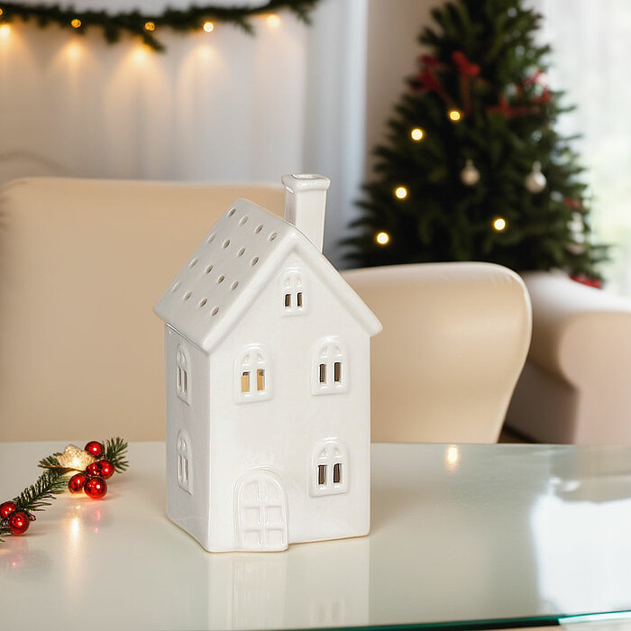 Ein weißes Keramikhaus mit kleinen beleuchteten Fenstern steht auf einem gläsernen Tisch. Daneben liegt ein Weihnachtsgesteck mit roten Beeren und grünen Zweigen. Im Hintergrund sind ein weißer Sessel und ein weihnachtlich geschmückter Baum mit leuchtende