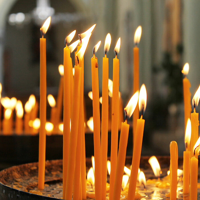 Das Bild zeigt eine Vielzahl brennender, langer, dünner Kerzen des Herstellers "Wenzel". Die Kerzen sind dicht an dicht in Metallschalen angeordnet, und ihre flackernden Flammen erzeugen ein warmes, feierliches Licht. Die Hintergrundkulisse ist leicht uns
