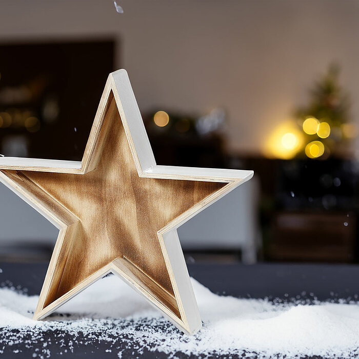 Ein Holzstern mit einer glänzenden, goldenen Innenseite steht auf einer dunklen Oberfläche. Um den Stern herum sind weiße Schneeflocken verstreut. Im verschwommenen Hintergrund sind dekorative Lichter und unscharfe weihnachtliche Dekorationen zu erkennen.