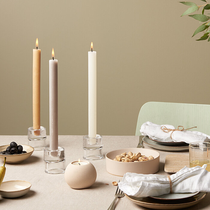 Eine lila Bolsius Rustikkerze liegt auf einem runden, schwarzen Tablett. Daneben befindet sich ein kleiner, schwarzer Kerzenhalter auf einer strukturierten, dunklen Unterlage.