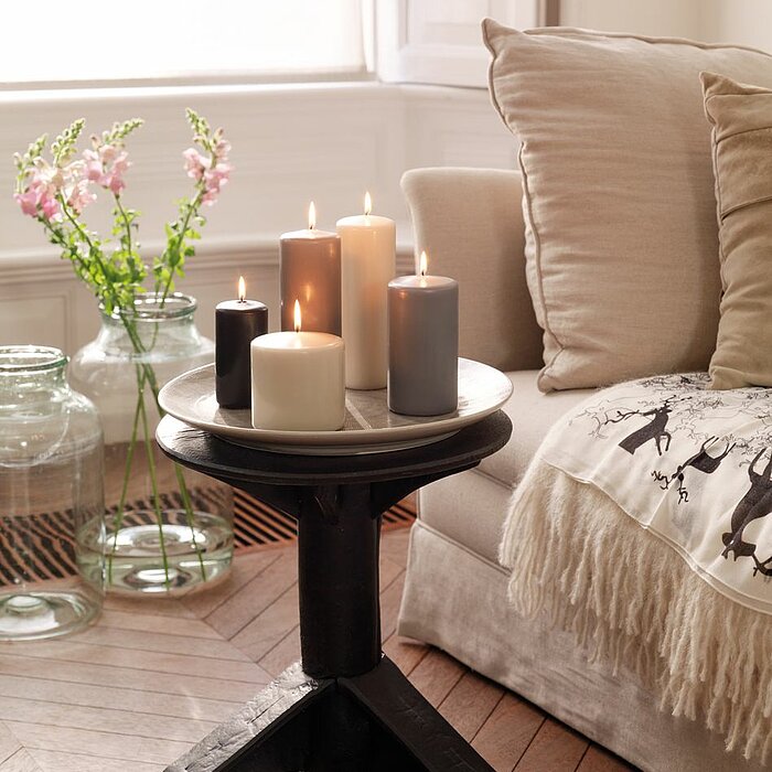 Vier brennende Bolsius Stumpenkerzen in den Farben Gelb, Orange und Rot stehen auf einem Keramikteller auf einem rustikalen Holztisch. Im Hintergrund befindet sich ein beiges Sofa mit blumigen und roten Kissen und einer roten Decke. Neben dem Keramiktelle