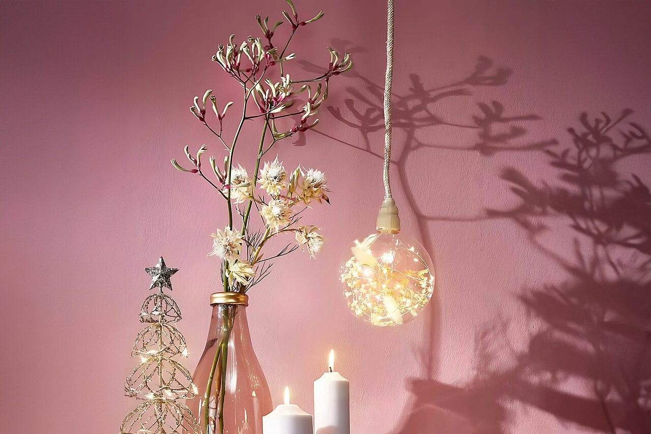 Vor einer altrosa Wand stehen verschiedene Dekorationsgegenstände, die einladend wirken: Ein filigraner Tannenbaum aus goldenem Draht, eine transparente Vase mit blühenden Zweigen, eine dekorative Glühbirne mit LED-Kette und getrockneten Blüten sowie zwei weiße Stumpenkerzen, die eine gemütliche Atmosphäre schaffen. Die Dekoobjekte werfen sanfte Schaffen auf die Wand.