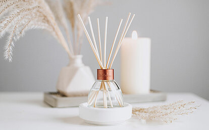 Ein freundlicher, heller Raum mit einer hellgrauen Wand. Auf einem weißen Tisch stehen chilf in einer weißen Vase und eine cremefarbene Stumpüenkerze im Hintergrund. Im Vordergrund ist ein eleganter Raumduft Diffuser mit Rattanstäbchen zu sehen.