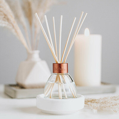 Ein freundlicher, heller Raum mit einer hellgrauen Wand. Auf einem weißen Tisch stehen chilf in einer weißen Vase und eine cremefarbene Stumpüenkerze im Hintergrund. Im Vordergrund ist ein eleganter Raumduft Diffuser mit Rattanstäbchen zu sehen.