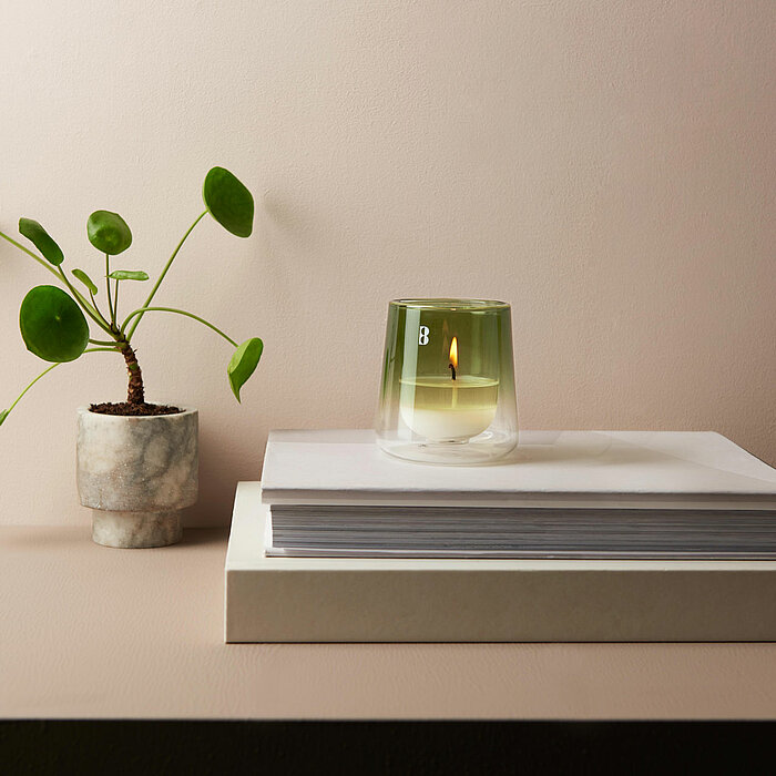 Eine brennende Bolsius Duftkerze in einem transparenten, hellgrünen Kerzenglas steht auf einem Stapel weißer Bücher. Neben der Duftkerze befindet sich eine kleine Pflanze im Topf. Der Hintergrund ist in einem sanften Beige gehalten.