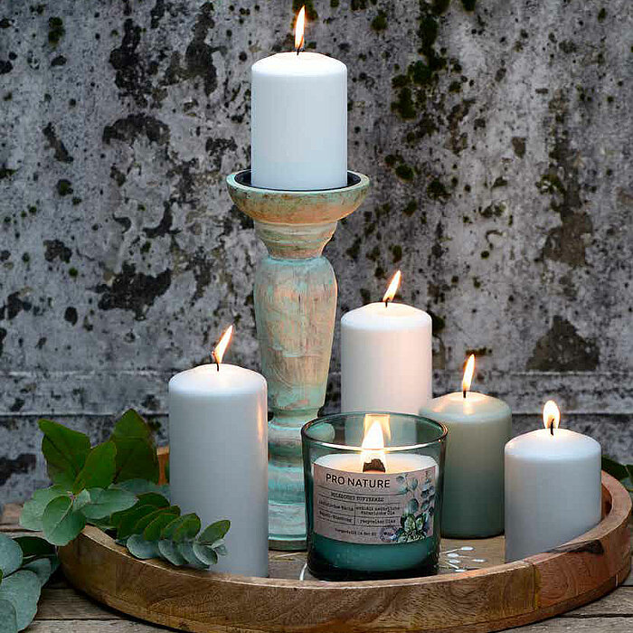 Zwei brennende weiße Stumpenkerzen und eine brennende Müller Duftkerze in einem grünen Glasbehälter mit der Aufschrift "Pro Nature" stehen gemeinsam auf einem Holztablett. Auf dem Tablett steht auch ein Blumentopf mit weißen Blüten und einer dekorativen M