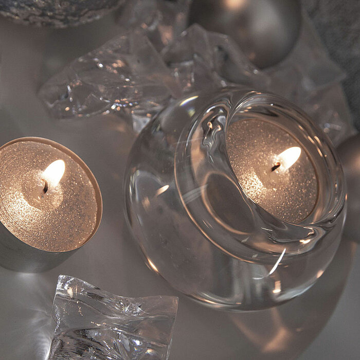 Zwei brennende Teelichter in silbernen Haltern stehen auf einem weißen Tablett. Um sie herum liegen durchsichtige, glänzende Dekorationselemente und zwei silberne Kugeln. Die sanft flackernden Flammen erzeugen eine gemütliche, festliche Atmosphäre.