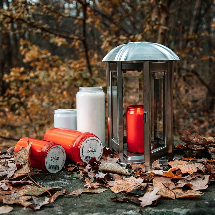 Vier nicht brennende Grablichter der Marke "DecoLite" stehen auf einem von Herbstlaub bedeckten Stein. Zwei rote und ein weißes Grablicht sind auf der linken Seite zu sehen, während ein weiteres rotes Grablicht in einer silbernen Laterne aufgestellt ist. 