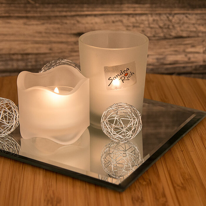 Ein brennendes Teelicht in einem matten, weiß gewellten Glasbehälter steht auf einer quadratischen Spiegelplatte. Daneben befindet sich ein gefrosteter Glasbehälter mit der Aufschrift "Sandra Rich". Um die Kerzen sind zwei silberne, filigrane Dekokugeln p