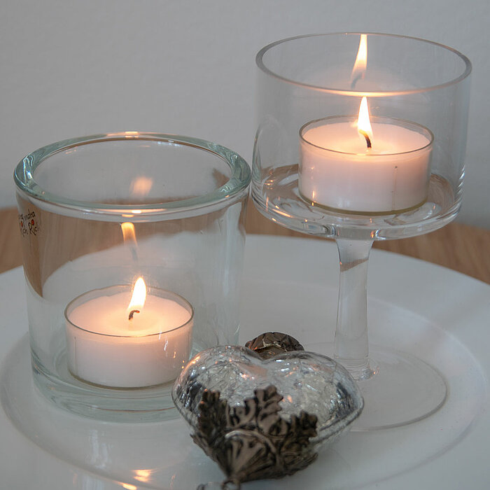 Zwei brennende Teelichter befinden sich in klaren Glasbehältern, die auf einem weißen Teller arrangiert sind. Ein Glasbehälter ist zylinderförmig, während der andere ein Weinglas ist. Vor den Teelichtern liegt eine kleine, verzierte Glasfigur. Der Hinterg