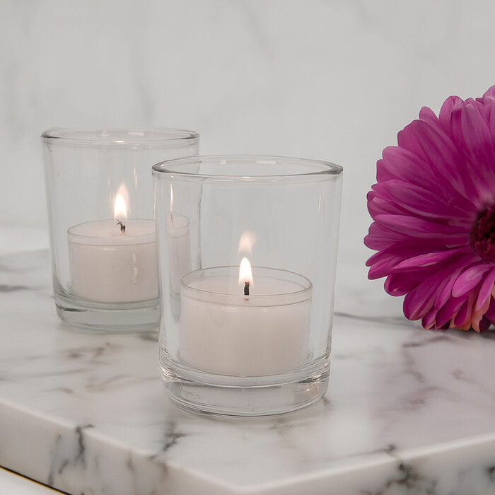 Zwei brennende Teelichter in Glasbehältern von DecoLite stehen auf einer marmorierten Fläche. Neben den Teelichtern liegt eine lilafarbene Blume. Die gesamte Szenerie vermittelt eine ruhige und elegante Stimmung.