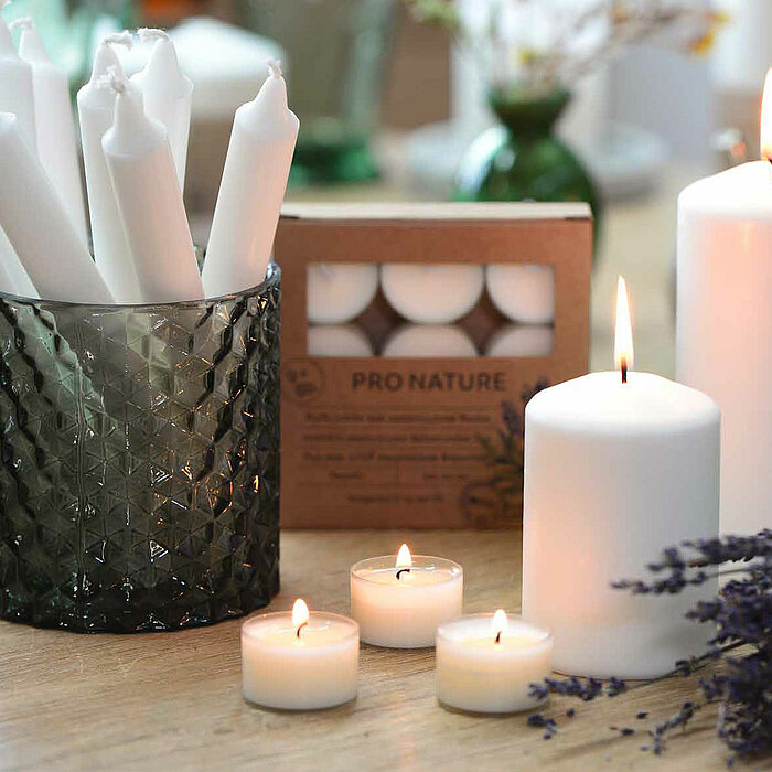 Vier brennende weiße Müller-Kerzen in verschiedenen Größen auf einem Holztisch, dekoriert mit Baumrinde und getrockneten Blumen. Eine der Kerzen befindet sich in einem filigran durchbrochenen weißen Kerzenhalter. Im Hintergrund steht ein grauer Topf mit l