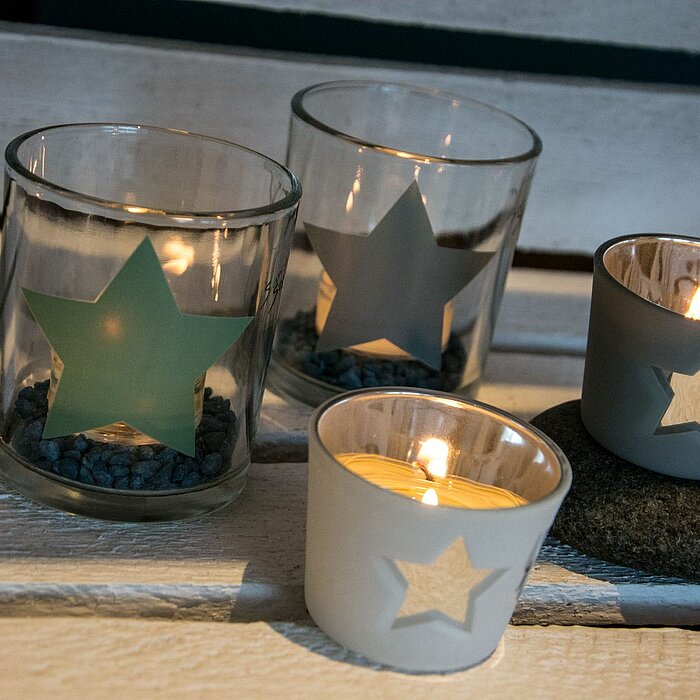 Sechs brennende Teelichter in farbigen Gläsern mit Sternmotiven stehen auf einem weißen Tablett. In der Mitte befindet sich eine größere grüne Laterne ebenfalls mit Sternmotiv vom Hersteller "Sandra Rich". Im Hintergrund sind Bücherregale und ein Sofa zu 