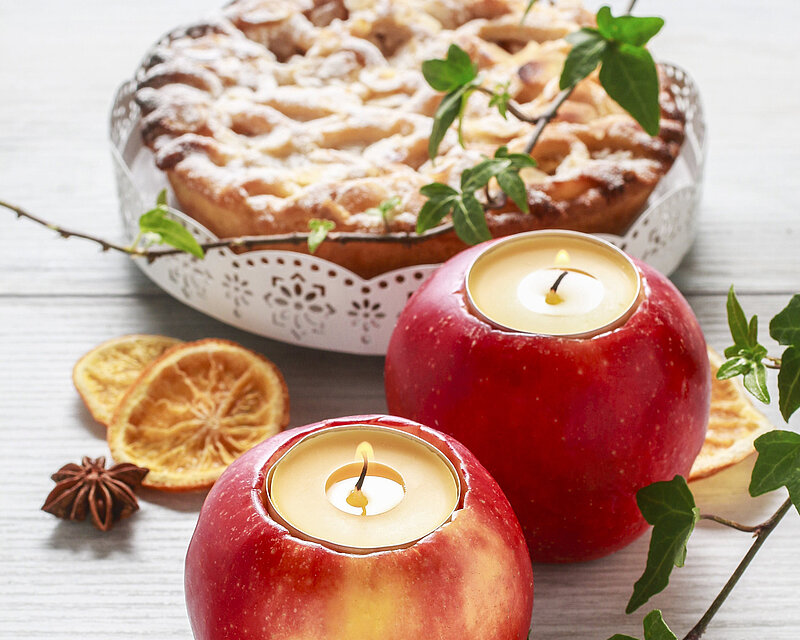 Auf einem weißen Holztisch, umgeben von zwei Efeu-Ranken, steht ein saftiger gedeckter Apfelkuchen neben zwei roten Äpfeln. Die Äpfel wurden eingeschnitten, und zwei Duft-Teelichter eingesetzt.