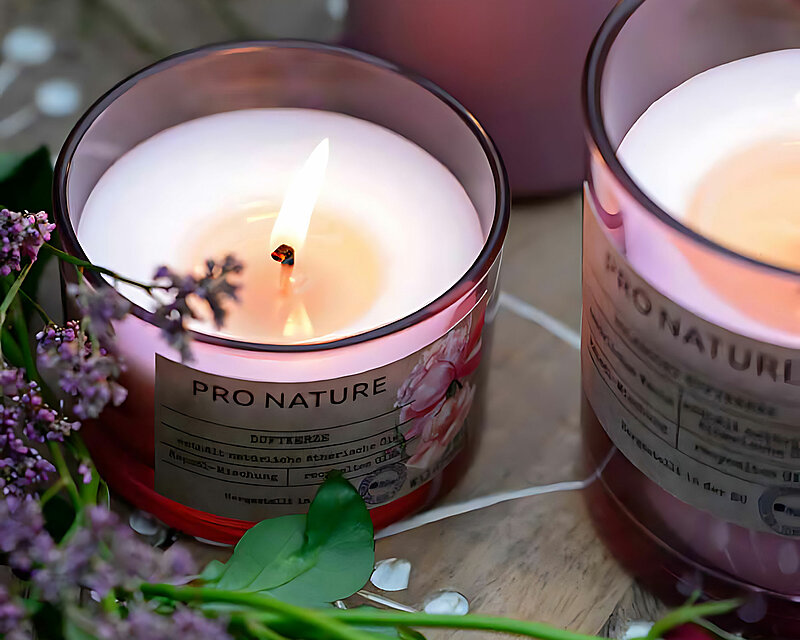 zwei brennende Rapsöl Duftkerzen im Glas mit dem Duft Wildrose stehen nebeneinander hinter einem Strauß verschiedenfarbiger rosen und Blüten. Im Hintergrund sieht man eine altrosafarbene Stumpenkerze