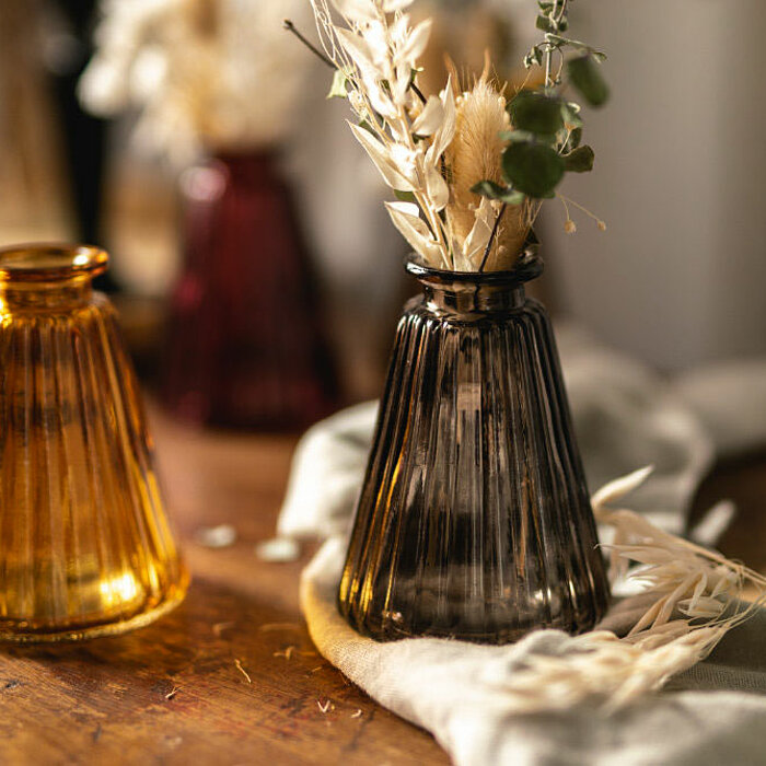 Die Produkte vom Hersteller "Sandra Rich" sind vier hübsche Glasvasen in verschiedenen Farben und Formen, die auf einem hölzernen Tisch arrangiert sind. Die Farben der Vasen sind warmes Orange, Rauchgrau, tiefes Grün und im Hintergrund ein intensives Rot.