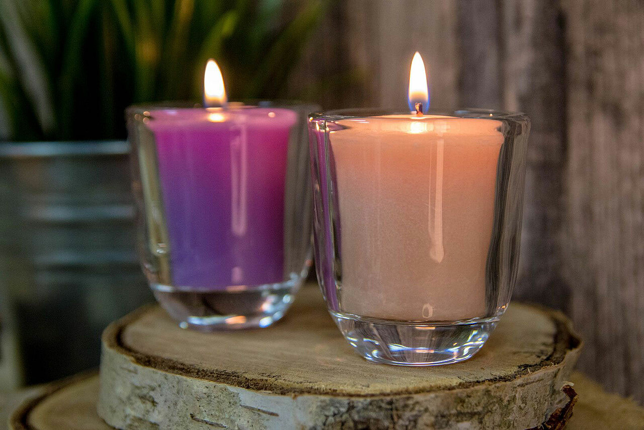 Das Bild hat eine gemütliche Ausstrahlung. Auf einer dicken Holzscheibe stehen zwei Votiv Gläser mit je einem lila und einem cremefarbenen brennenden Votiv. Im Hintergrund erkennt man eine Zinkschale mit einer grünen Pflanze.
