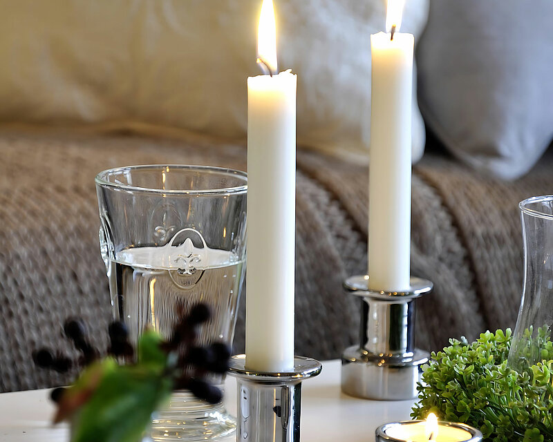 Vor einem gemütlichen Sofa steht ein weißer Couchtisch. Auf dem Couchtisch stehen zwei silberne Stabkerzenhalter mit weißen brennenden Stabkerzen- Daneben steht ein silberner Teelichthalter mit einem brennenden Teelicht. Auf dem Tisch steht ein Trinkglas gefüllt mit Wasser und daneben stehen mehrere Glasvasen mit grünen zweigen und beeren.