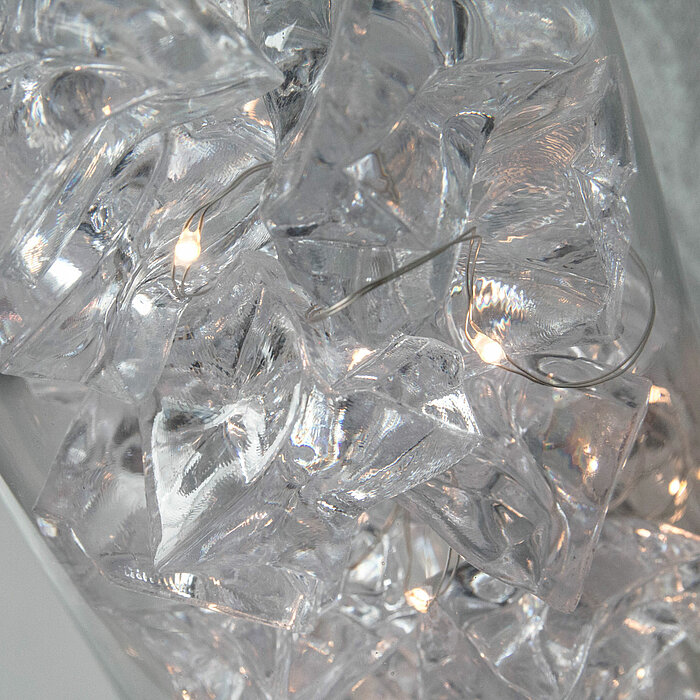 Dicht gepackte, durchsichtige Kunststoff- oder Glassteine, die mit einer Lichterkette durchzogen sind. Die Steine und die kleinen, warmweiß leuchtenden Lämpchen erzeugen ein funkelndes und glitzerndes Erscheinungsbild.