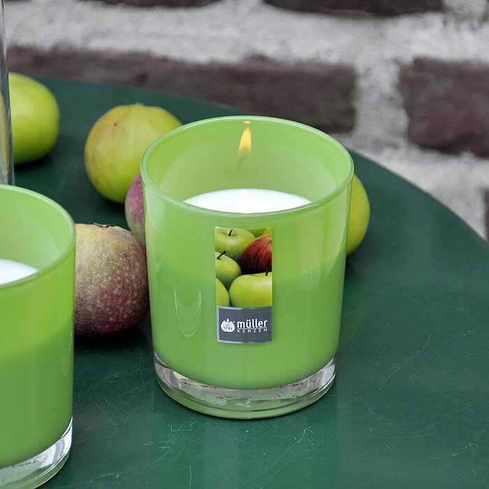 Zwei brennende grüne Duftkerzen des Herstellers "Müller" auf einem runden grünen Tisch. Im Hintergrund liegen mehrere grüne Äpfel sowie ein hohes Glasgefäß mit weißen Dekosteinen. Die Stimmung wirkt frisch und natürlich.