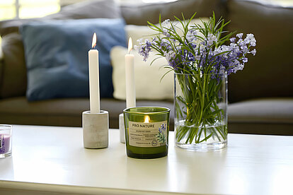 Auf dem Bild ist im Hintergrund eine gemütliche Couch zu sehen. Im Vordergrund steht ein weißer Wohnzimmertisch. Auf dem Tisch befindet sich ein Pro Nature Rosmarin-Lavendel Duftglas von Müller Kerzenfabrik, daneben zwei Betonhaltern von Müller Kerzenfabrik, in denen jeweils eine brennende weiße Stabkerze von Müller Kerzenfabrik steckt. Neben den Kerzen steht eine Glasvase, gefüllt mit etwas Wasser und einem Bündel Rosmarin und Lavendel. Das Zimmer ist hell und lichtdurchflutet, was eine frische und entspannte Atmosphäre schafft.