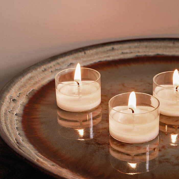 Drei brennende Bolsius Teelichter stehen auf einem großen, rustikalen Keramiktablett mit einer glänzenden Oberfläche. Die Teelichter verbreiten ein warmes, sanftes Licht in der Umgebung.